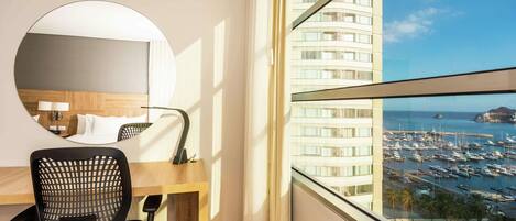 A hotel room with a large window, a round mirror, a wooden desk, and a black office chair.