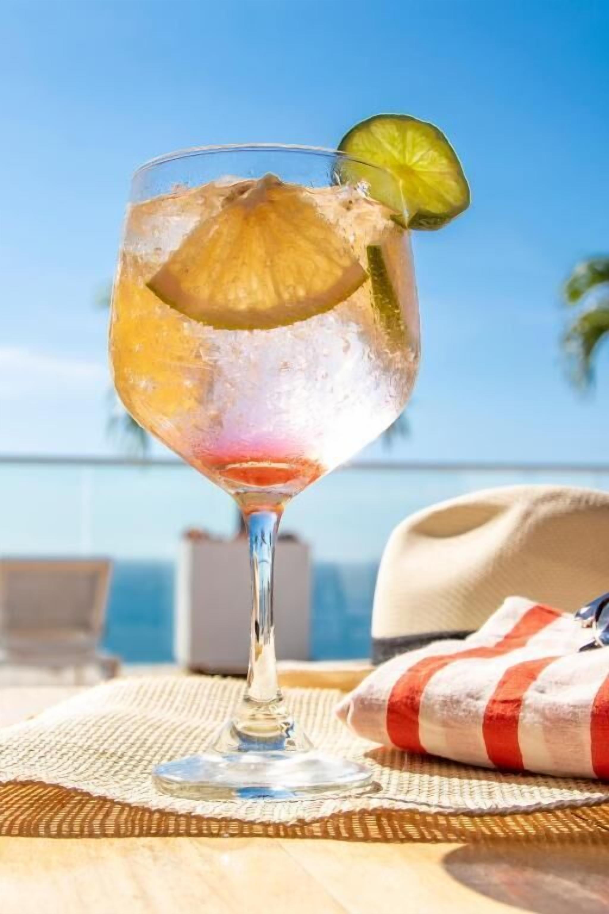 Un verre à cocktail, garni d’une tranche de citron vert, est posé sur une table recouverte d’un set de table en fibres tressées, d’une nappe à rayures, et un chapeau est visible à l’arrière-plan.