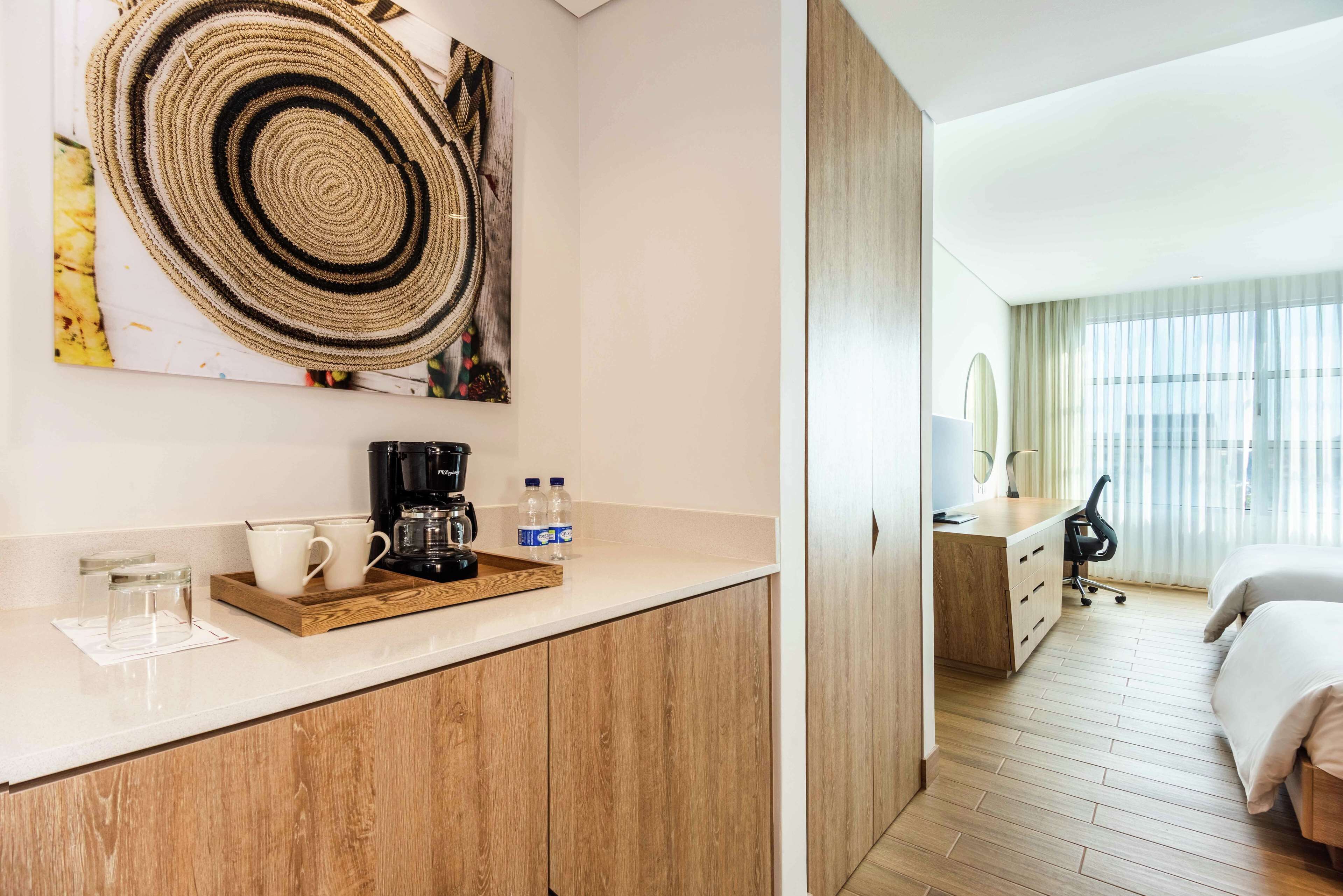 A hotel room with a wooden floor, a bed, a desk with a chair, a coffee maker, and a large window with curtains.