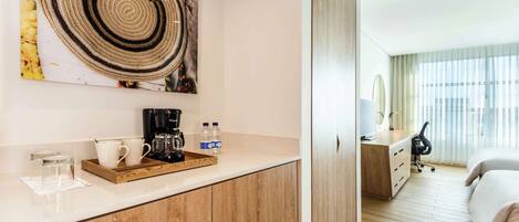 A hotel room with a wooden floor, a bed, a desk with a chair, a coffee maker, and a large window with curtains.