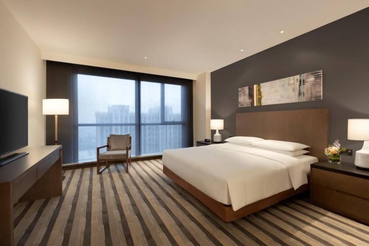 Een hotelkamer met een groot bed, een bureau, een stoel, een televisie en uitzicht op de stad door grote ramen.