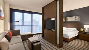 Hotelkamer met groot raam, flatscreen televisie, bed met witte lakens, sofa met meerdere kussens en een kleine tafel met lamp.