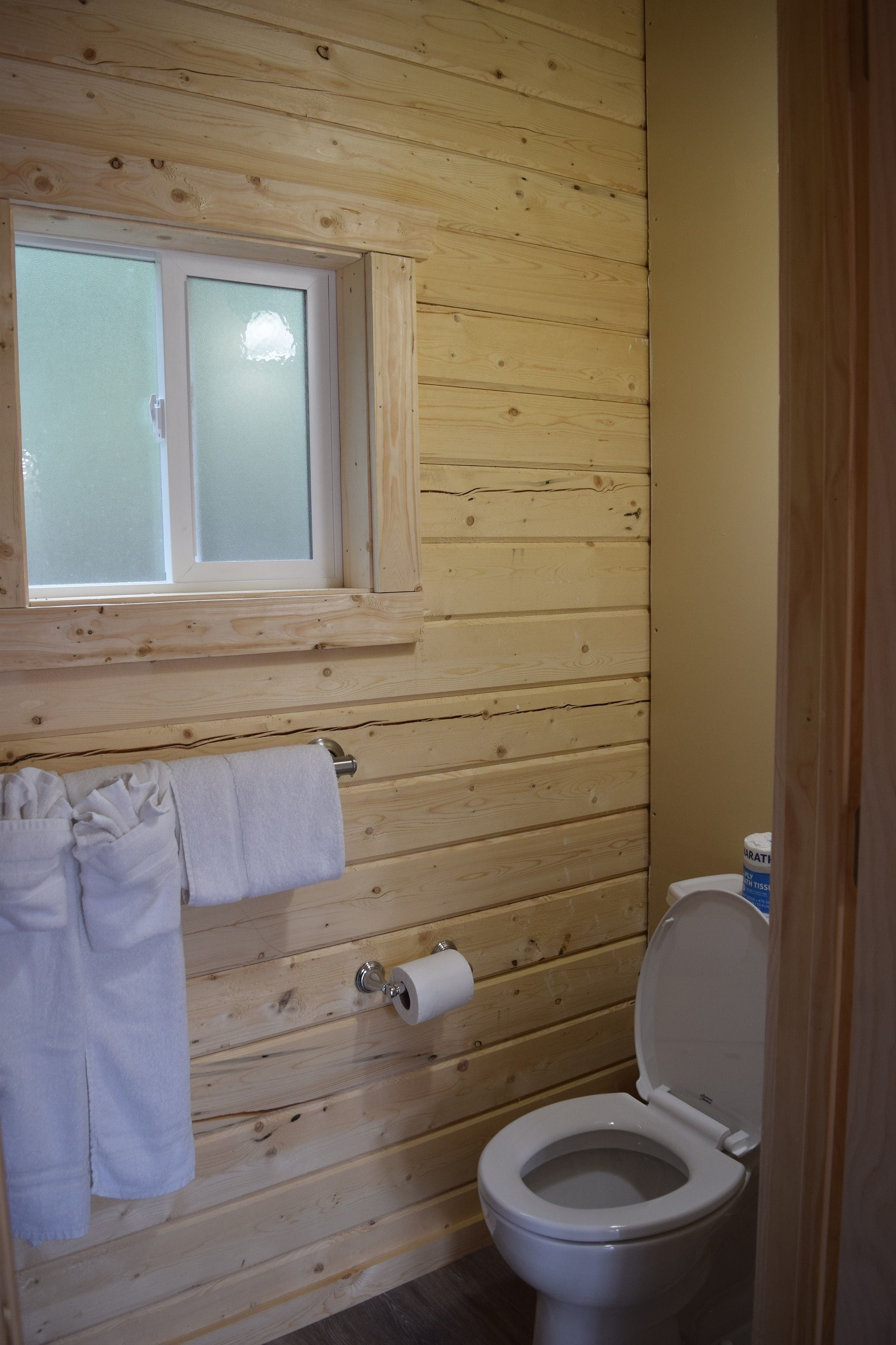 A wooden-paneled bathroom with a toilet, a window, and a towel rack.