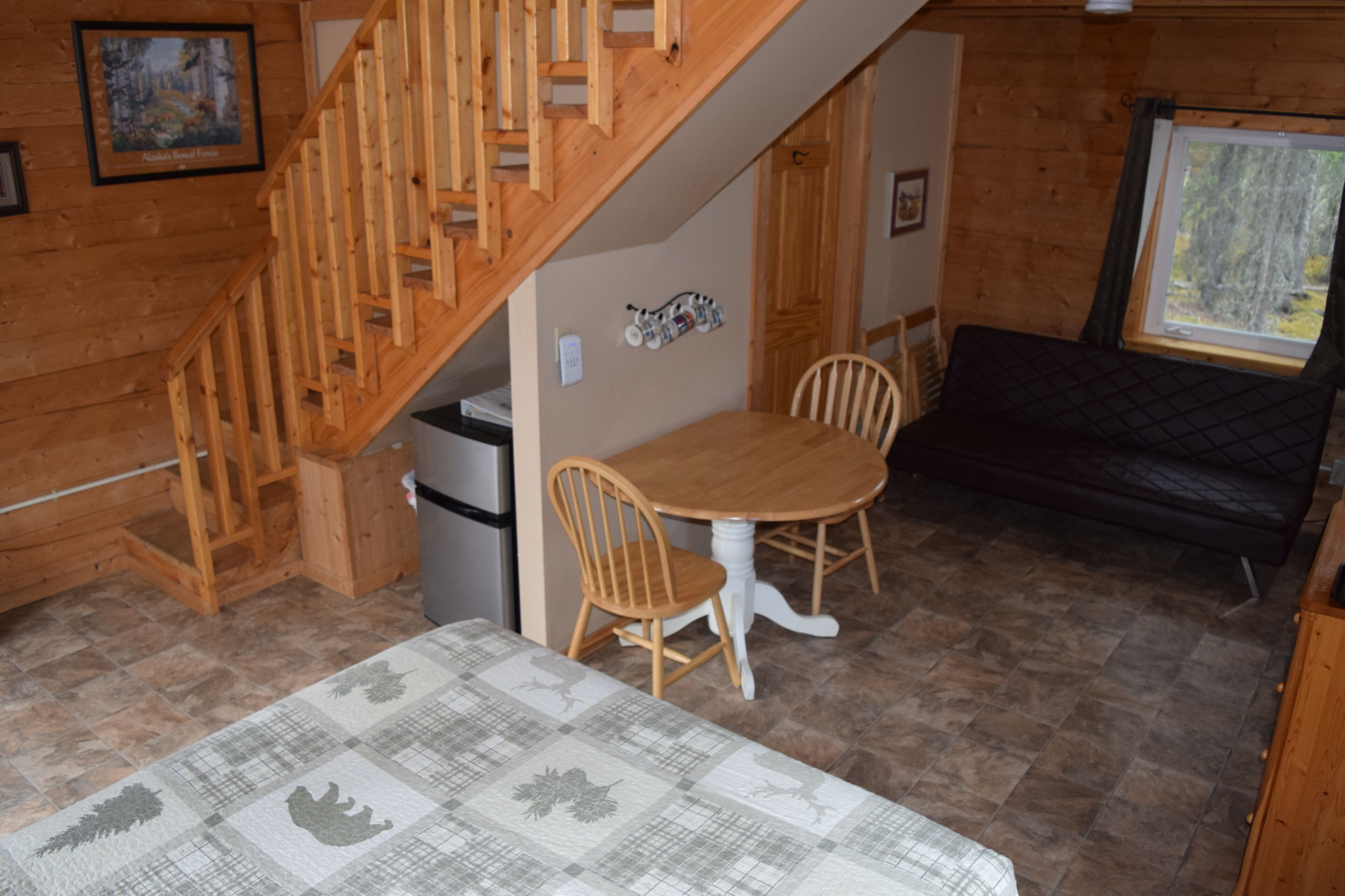 A cozy cabin interior with a wooden staircase, a dining table with chairs, a sofa, and a window with a view of trees.
