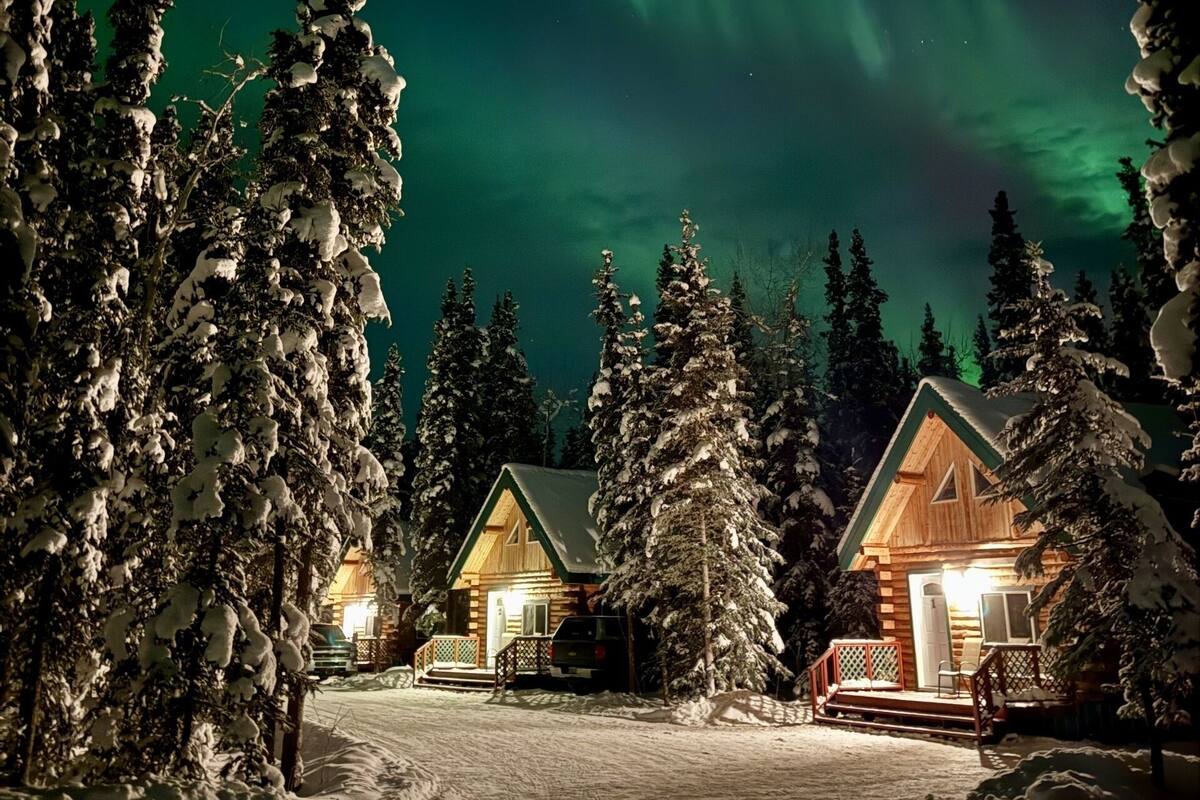 A cabin with a snow-covered landscape, surrounded by trees, under a night sky with aurora borealis.