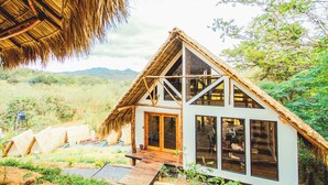 A thatched-roof house with a wooden deck and large windows, surrounded by greenery and hills. - Socialtel Maderas (San Juan del Sur)