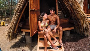 A couple sitting on a bench outside a thatched-roof structure with a wooden door, surrounded by trees and a clear sky. - Socialtel Maderas (San Juan del Sur)