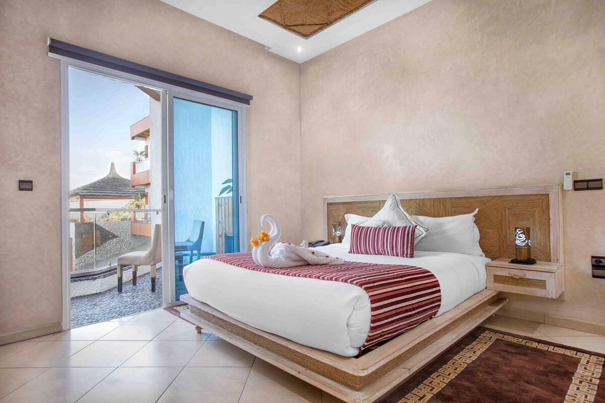 A hotel room with a large bed, a nightstand, a bedside table, a sliding glass door to a balcony, and a view of a building outside.