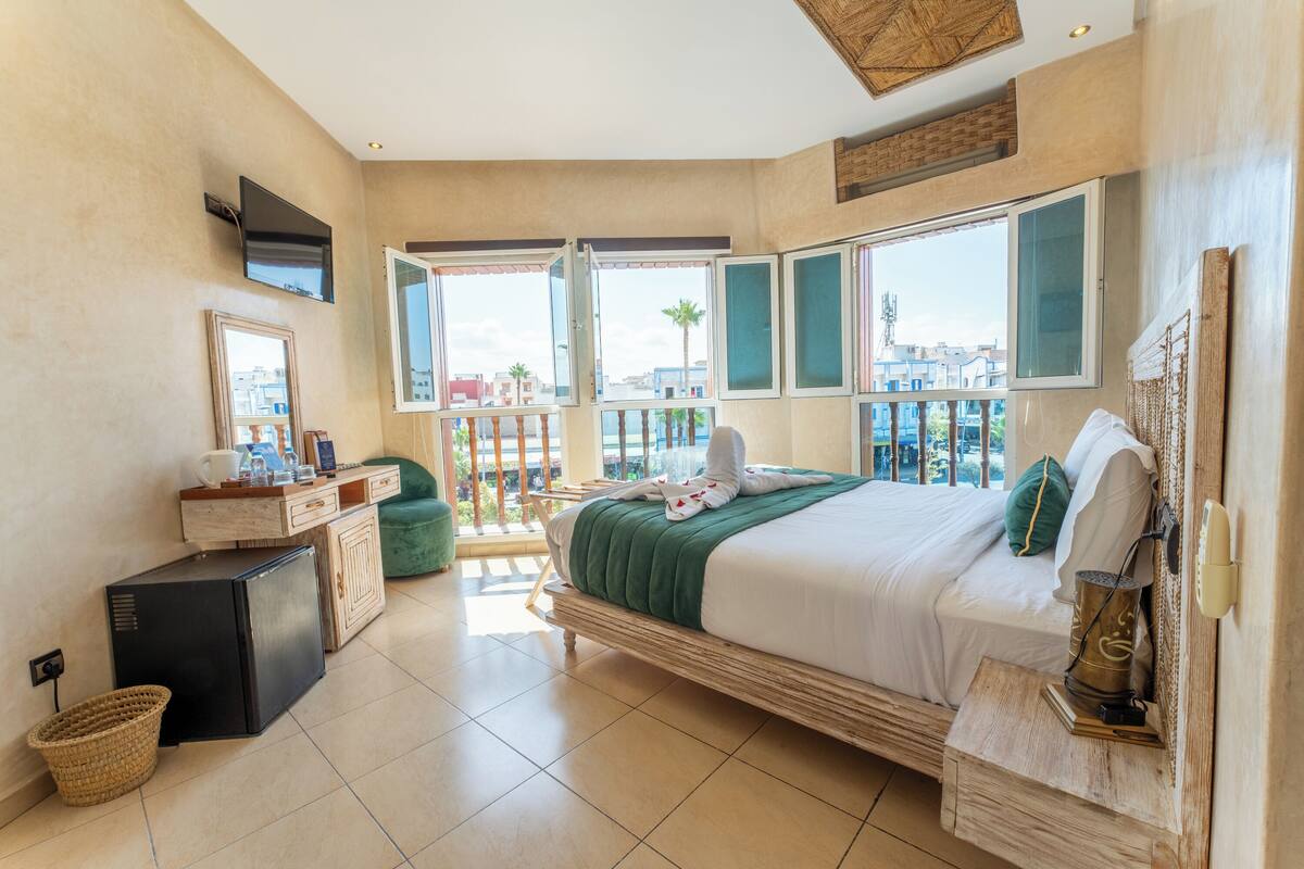 A hotel room with a bed, a desk, a television, and a balcony with a view of buildings and palm trees.