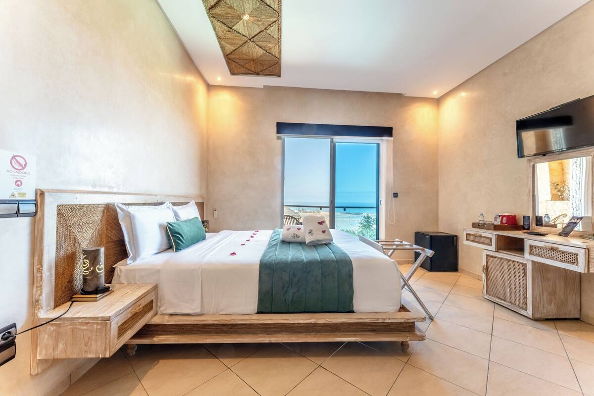 A modern hotel room with a large bed, a TV, a minibar, and a balcony with a view.