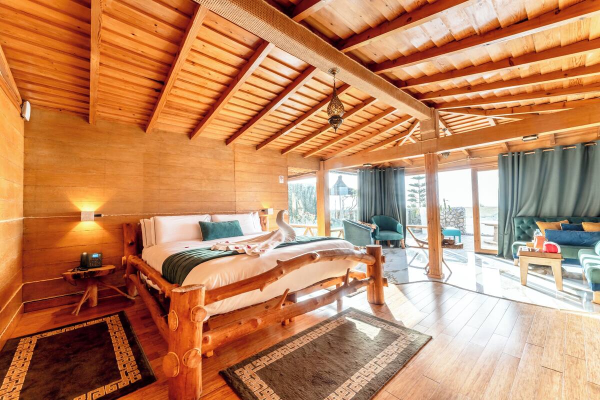 A wooden cabin with a large bed, a small table, and a chair. There is a window with curtains and a view of the outside.