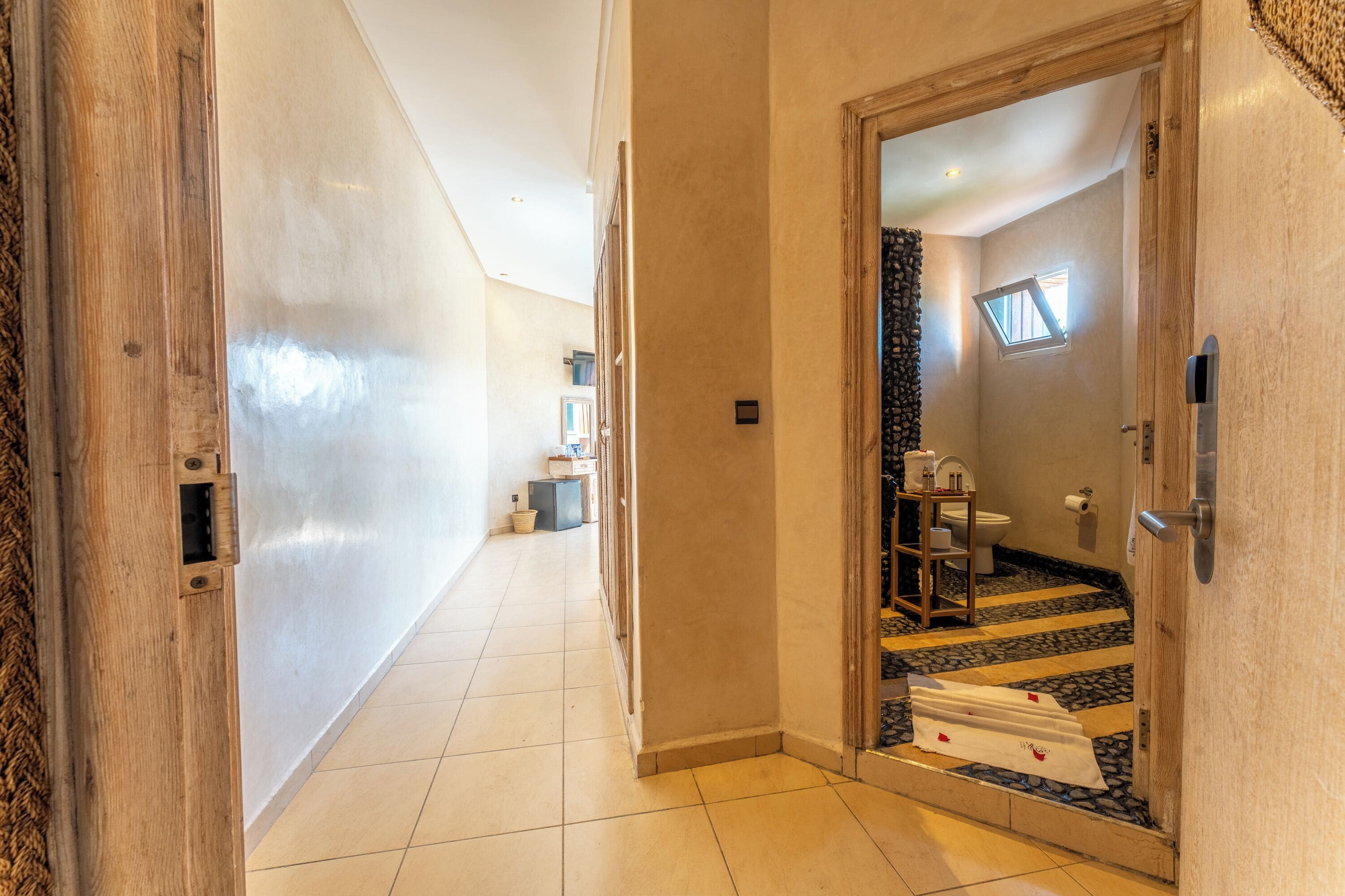 A hallway leading to a bathroom with a tiled floor, a wooden door, and a small window.