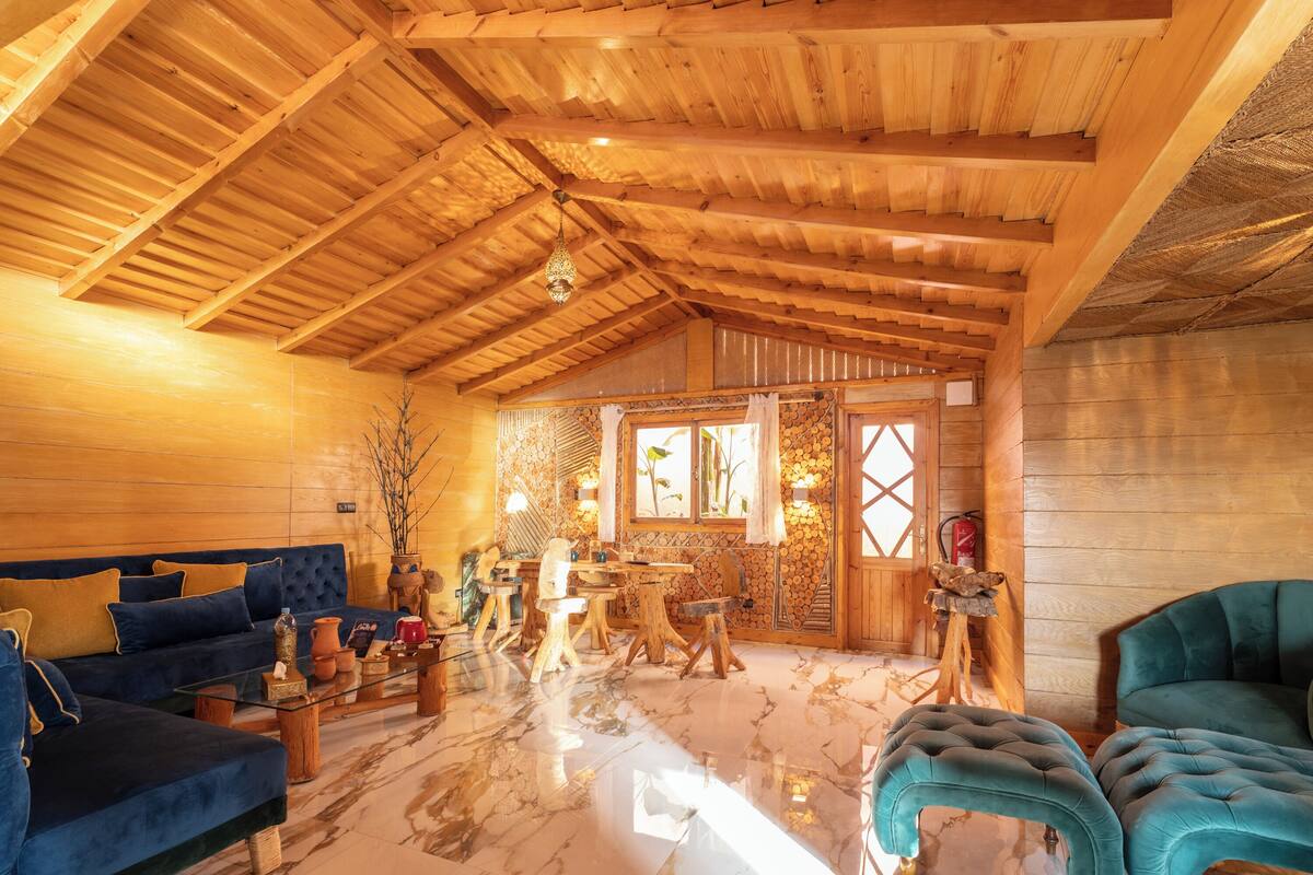 A spacious room with a high wooden ceiling, a marble floor, a blue sofa, a teal armchair, and a wooden dining table.