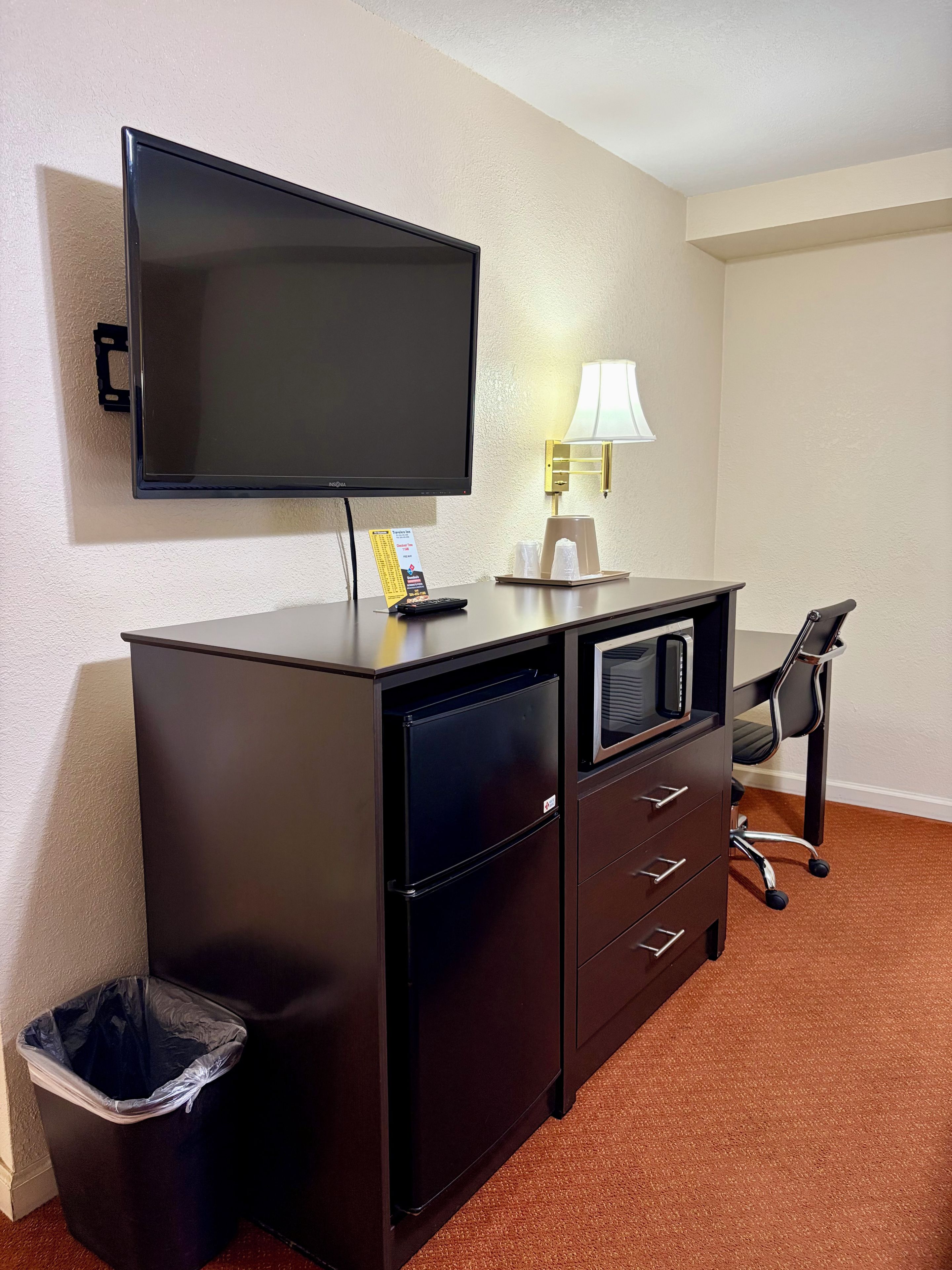 A hotel room with a flat-screen TV mounted on the wall, a desk with a lamp, a trash bin, and a chair.