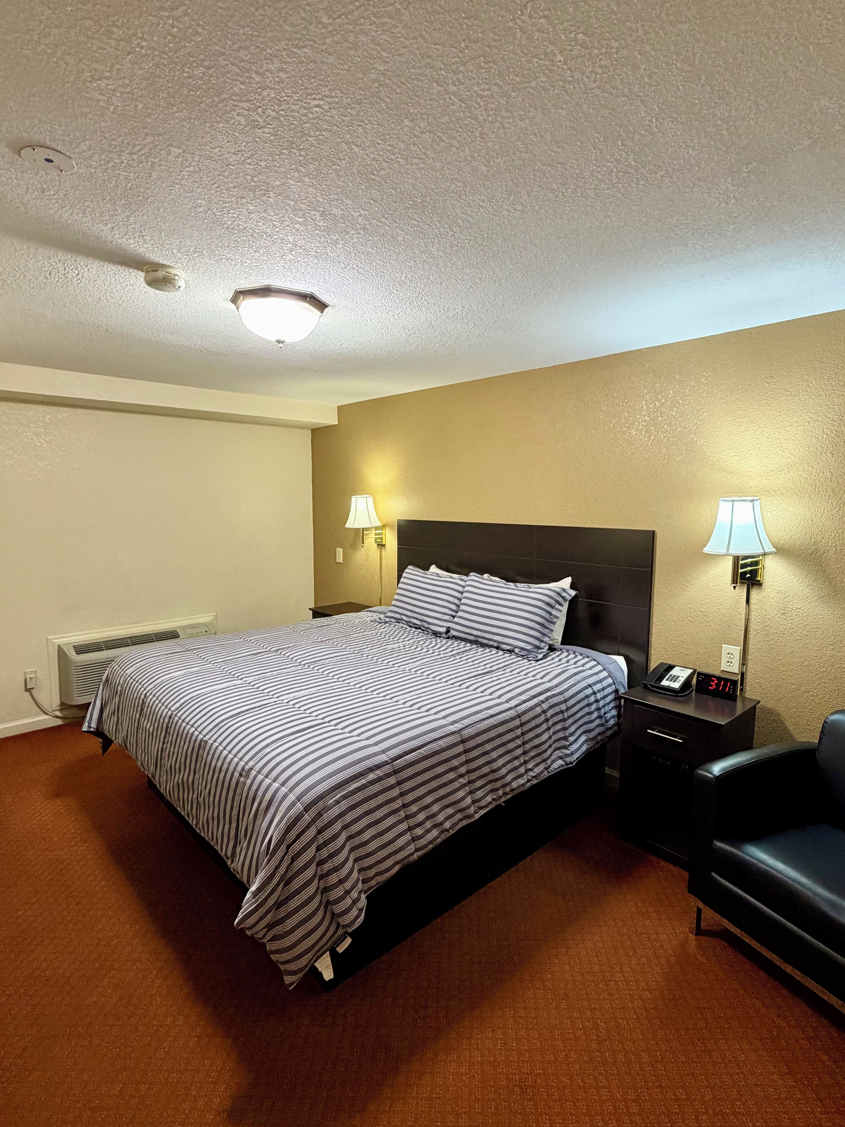 Habitación de hotel con cama, lámparas de noche, una silla, mesita de noche con reloj y un calentador de pared.