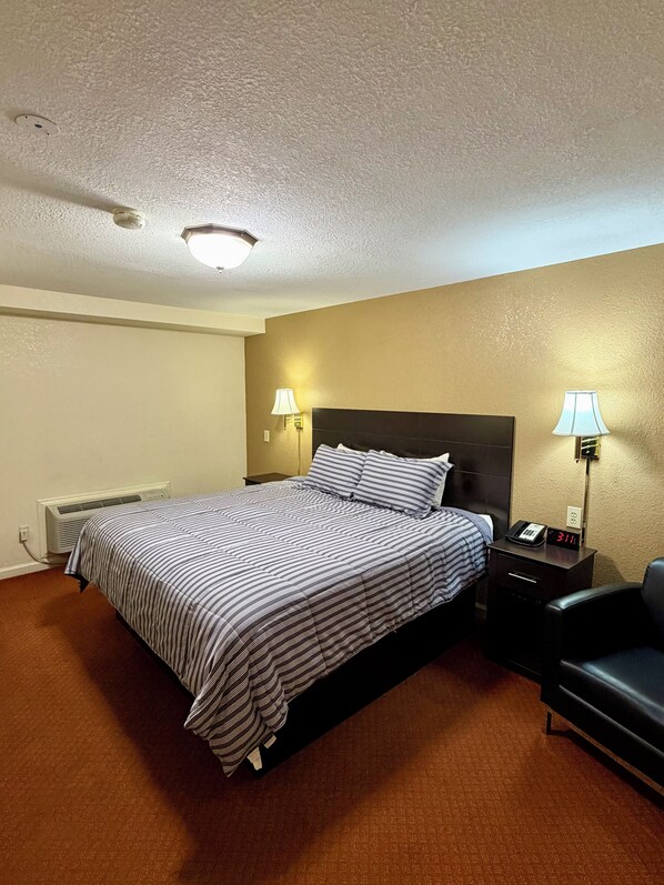 A hotel room with a bed, bedside lamps, a chair, a nightstand with a clock, and a wall-mounted heater.