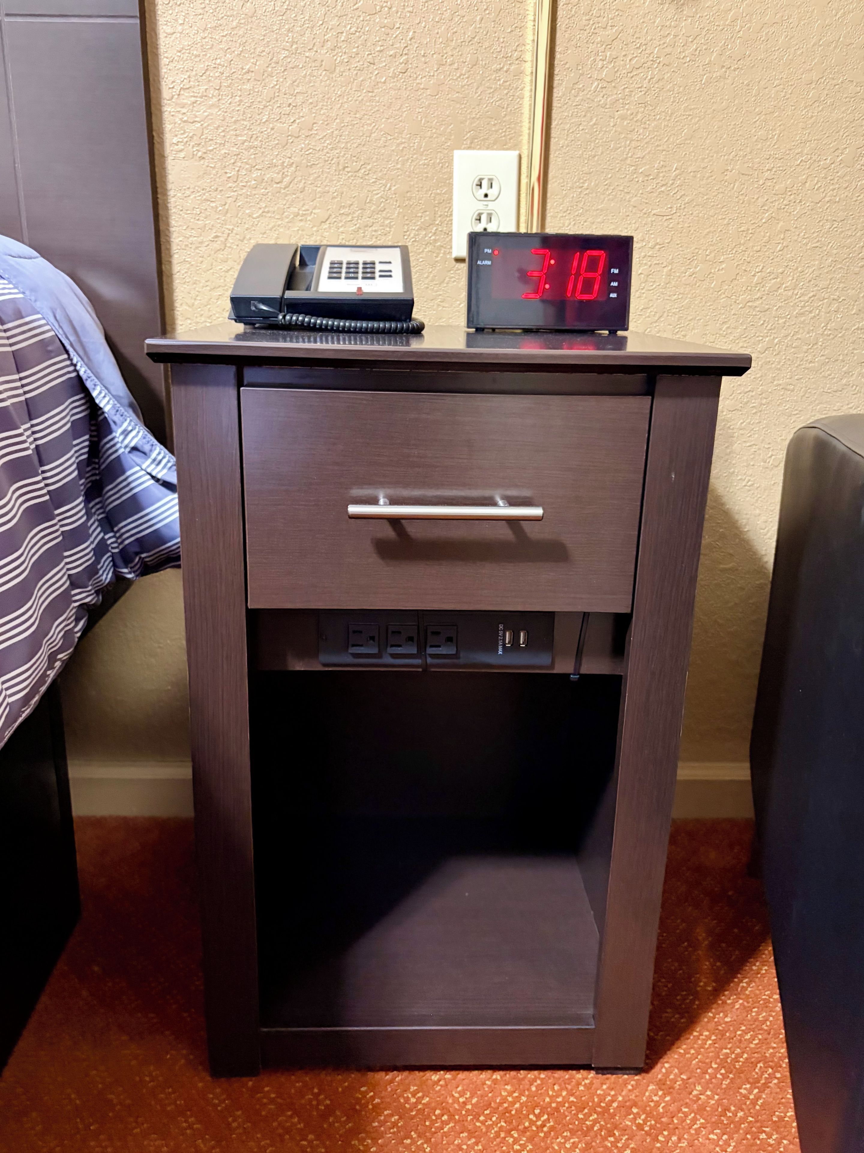 A wooden bedside table with a digital clock, a telephone, and a power outlet.