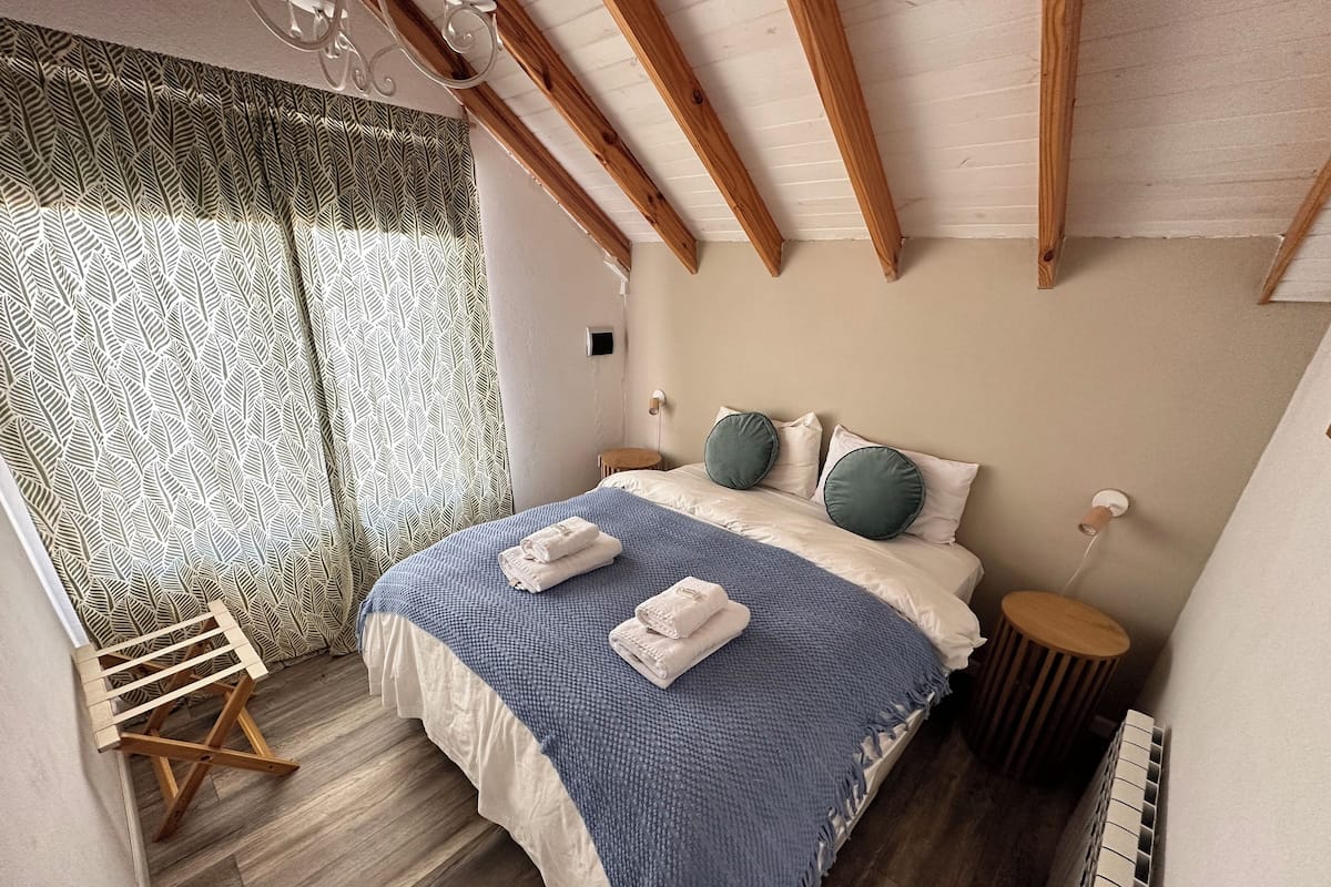 A bedroom with a bed, wooden beams, a radiator, and a window with patterned curtains.