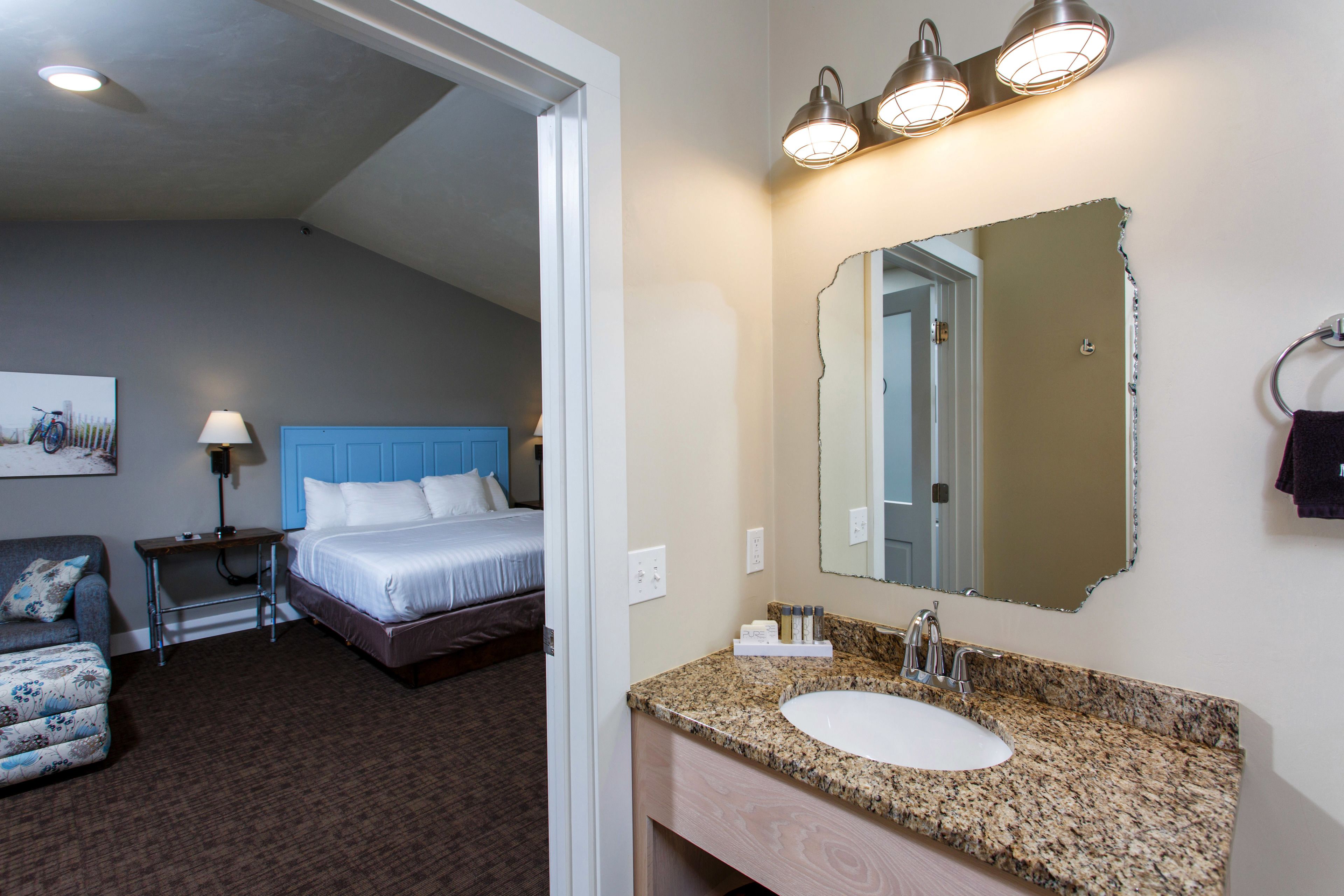 A hotel room with a bed, a bedside table, a chair, a bathroom with a sink and mirror, and a wall-mounted light fixture.