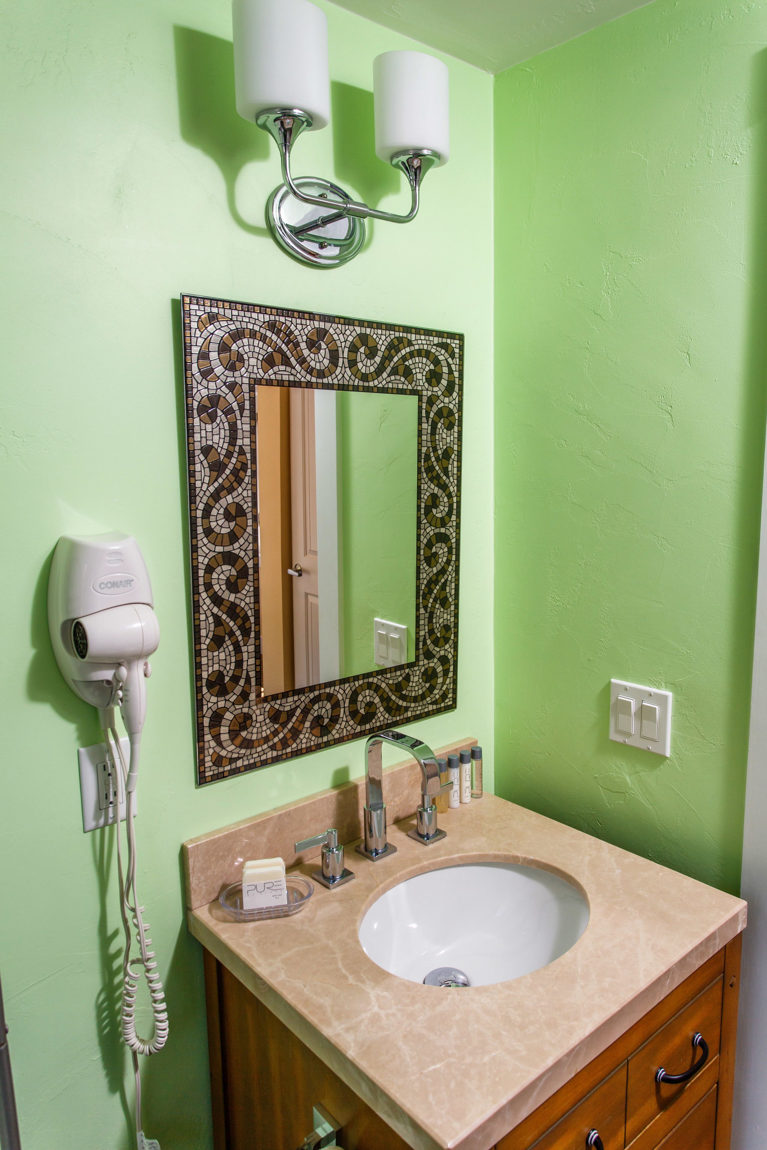 A bathroom with a green wall, a white sink, a mirror with a mosaic frame, a hairdryer, and a light fixture.