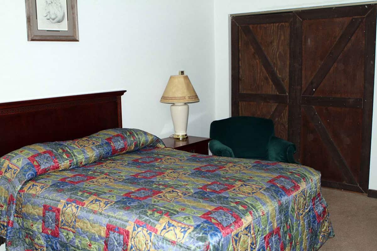 A bed with a colorful quilt, a bedside lamp, a chair, and a wooden door.