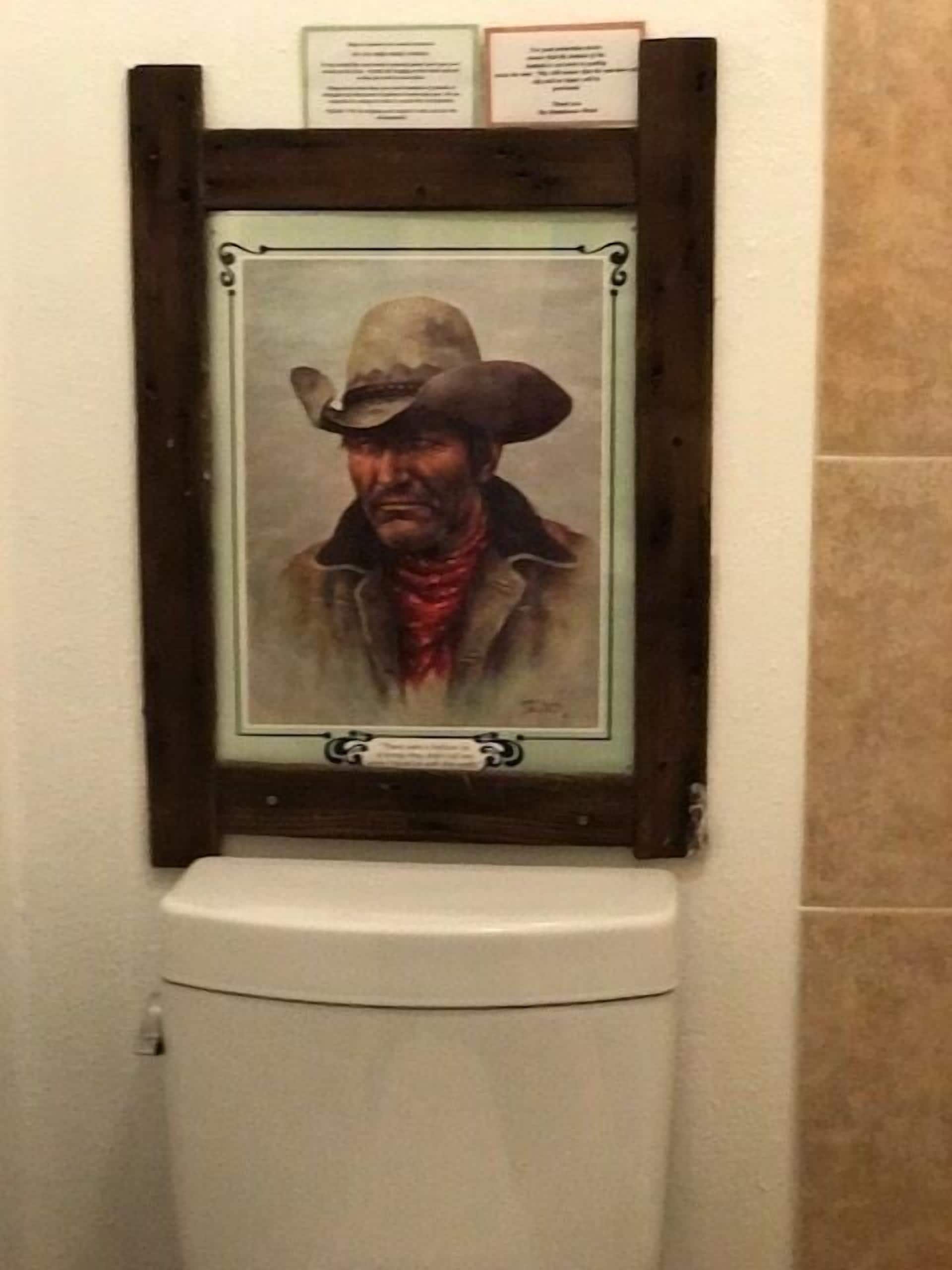 A framed portrait of a person in a cowboy hat and coat hanging on a wall above a toilet.