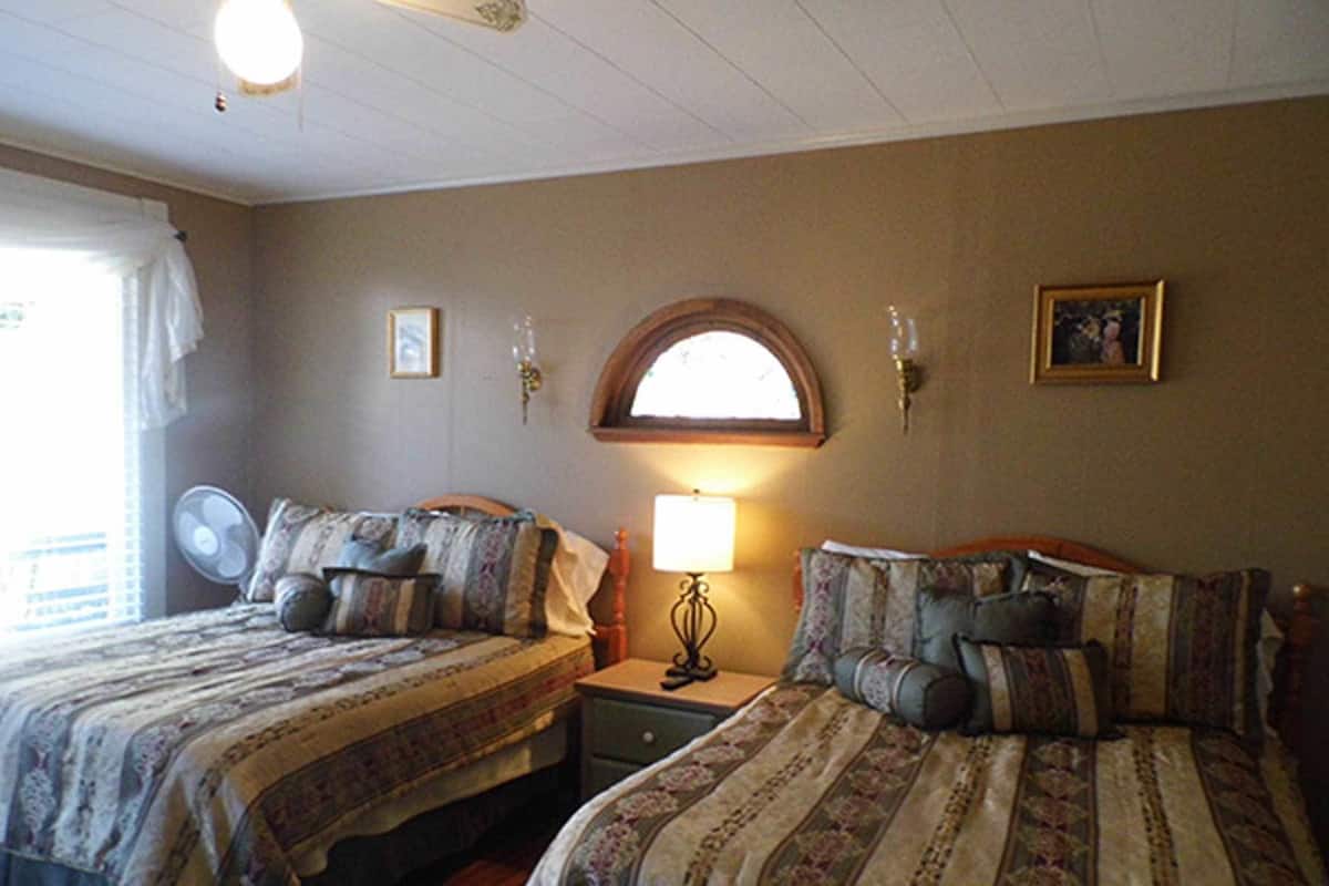 A room with two beds, a nightstand with a lamp, a wall-mounted clock, and a window with curtains.