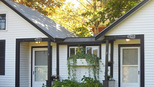 A white house with a black roof, two doors, and a small porch area with greenery. - Otter Creek Inn (Mount Desert)
