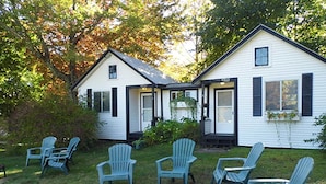 A two-story white house with black shutters, a front porch, and a small garden with chairs. - Otter Creek Inn (Mount Desert)