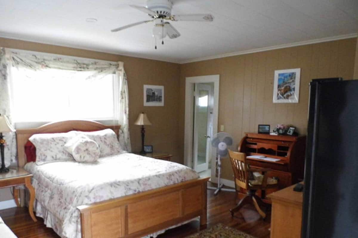 A bedroom with a wooden bed, a desk, a chair, a ceiling fan, and a door.
