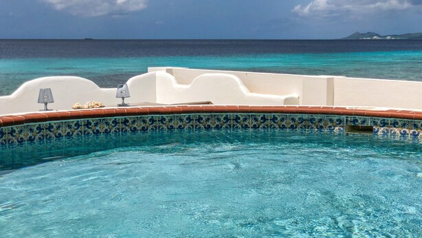 Una piscina con agua azul cristalina, borde de azulejos y vista al océano y la costa distante.