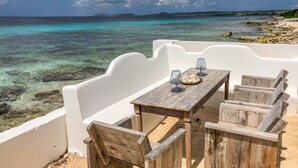 Una mesa de madera con sillas en un balcón con vista al mar turquesa, aguas cristalinas y costa rocosa.
