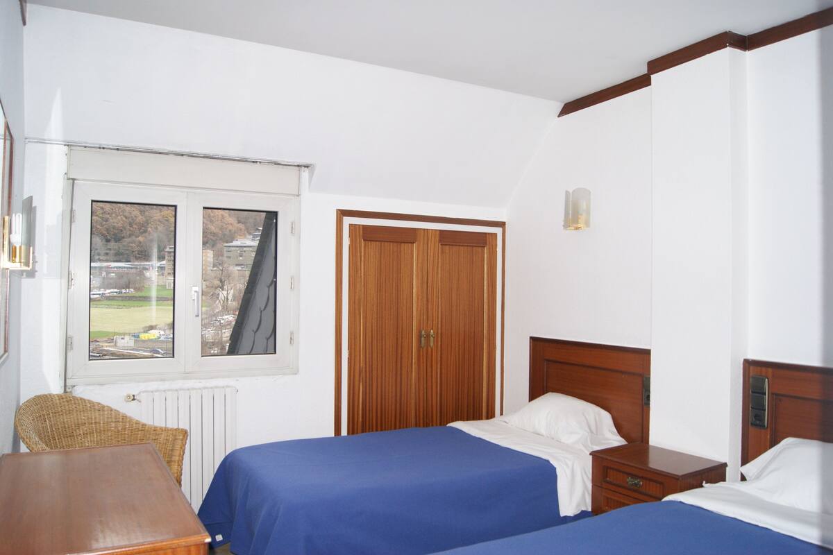 A bedroom with two beds, a wooden wardrobe, a window with a view of the outside, and a small table.