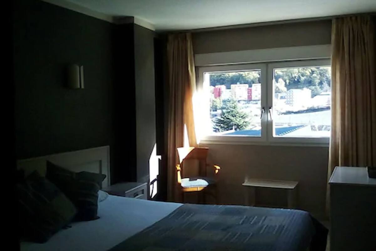 A hotel room with a bed, a chair, a small table, and a window with curtains.