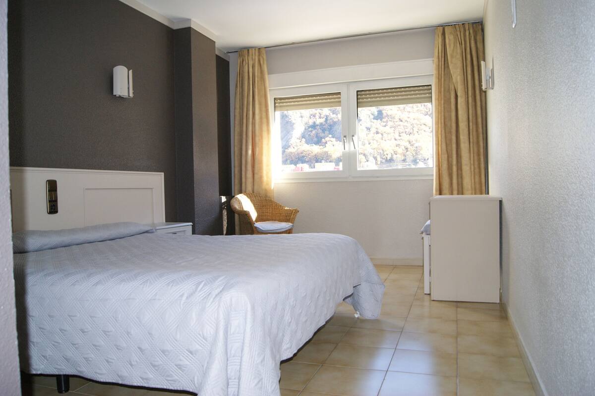 A hotel room with a bed, a chair, a small table, a window with curtains, and a refrigerator.