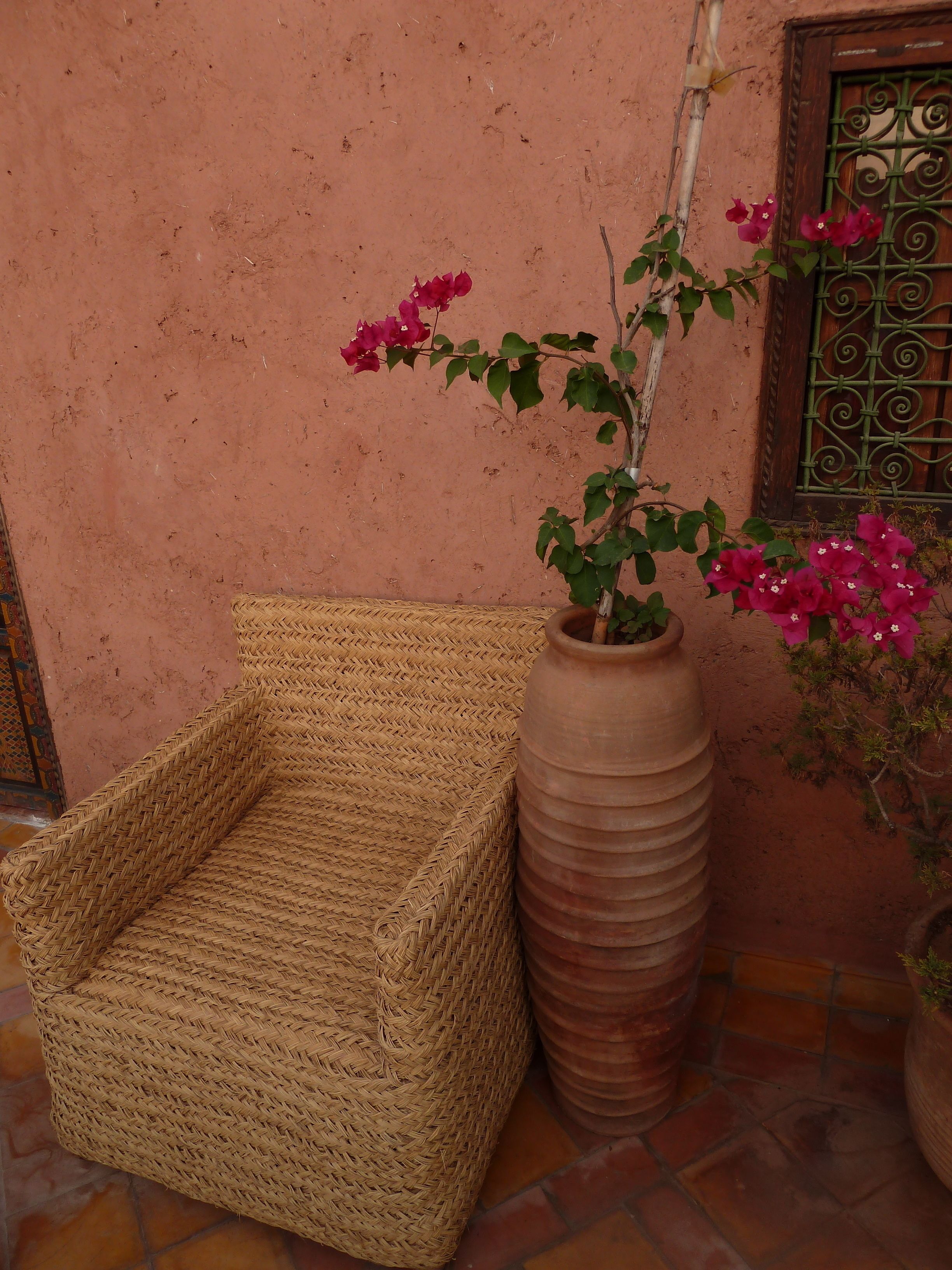 A wicker chair and a large terracotta pot with bougainvillea flowers on a tiled floor.