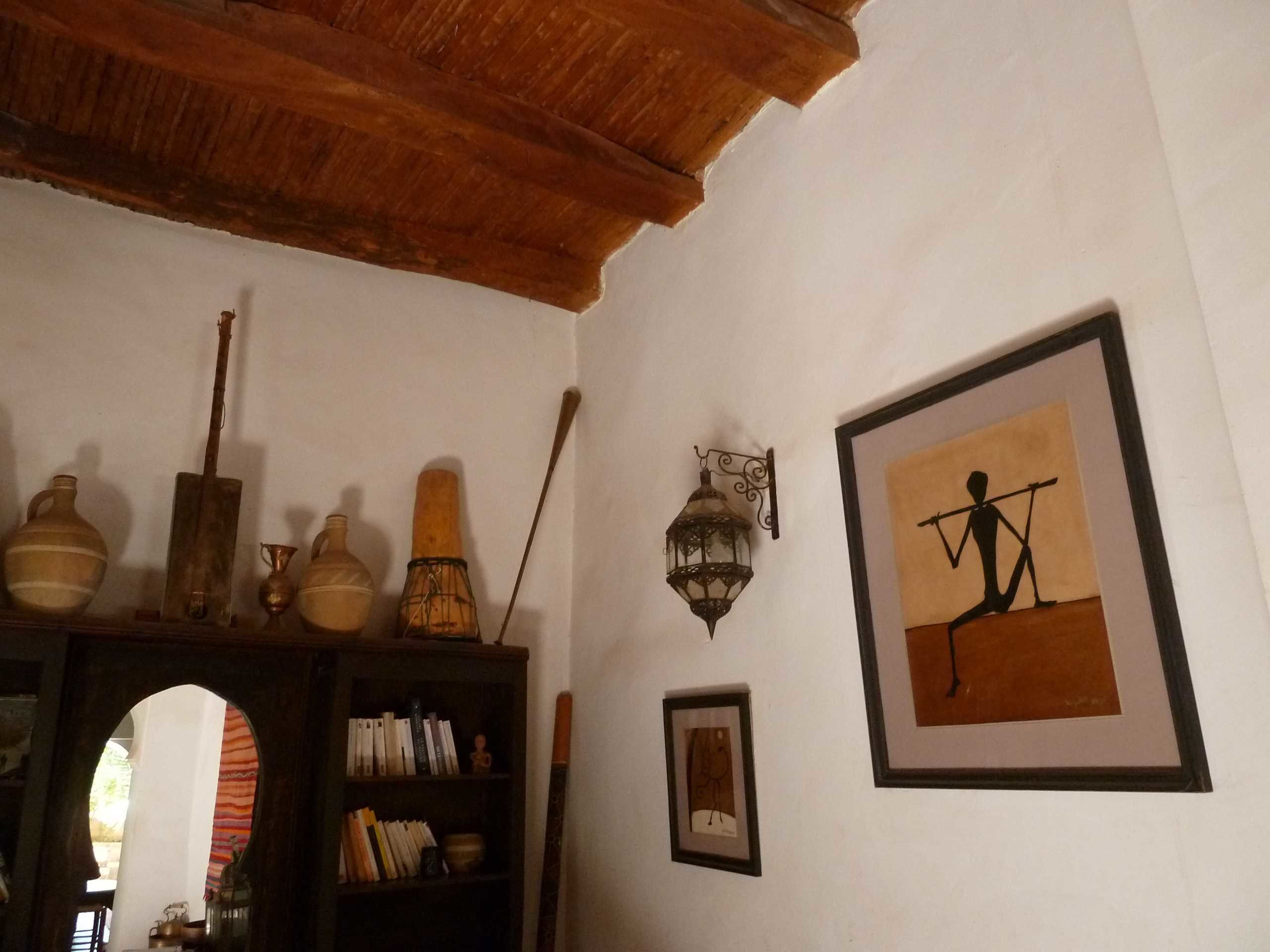 Una habitación con techo de madera, una estantería con libros y adornos, un cuadro enmarcado de una figura sosteniendo un asta, y un farol colgante en la pared.