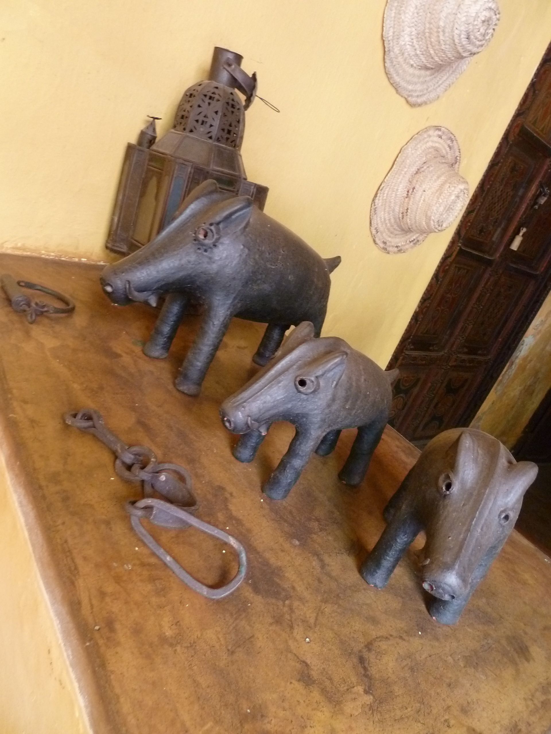 A rustic interior with three pig sculptures on a wooden surface, a lantern, and woven hats on the wall.