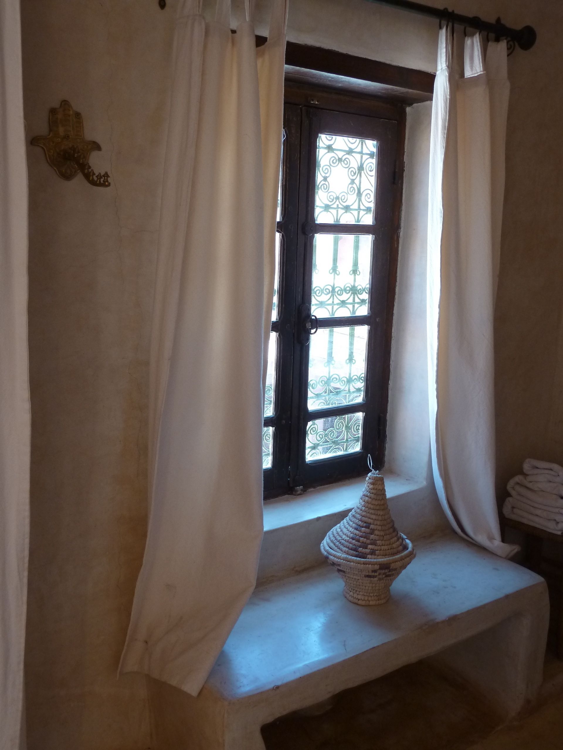 Ein Zimmer mit Fenster, weißen Vorhängen, einer kleinen Bank und einem gewebten Korb.