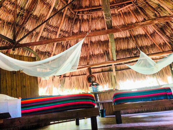 Una cabaña con techo de paja que tiene dos camas, cada una con ropa de cama de colores, y un dosel colgante.