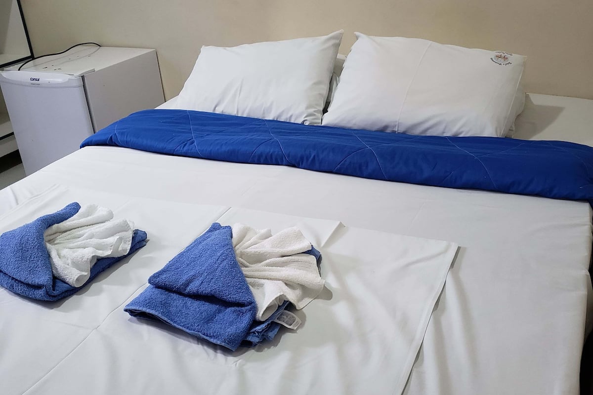 Una cama bien hecha con ropa de cama blanca y azul, una almohada blanca con un logotipo y una mesita de noche blanca con un distribuidor de corriente.