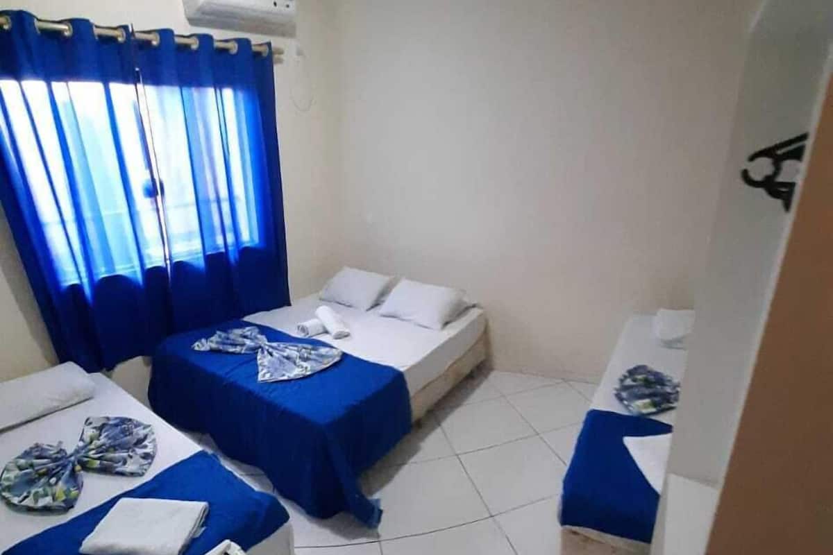 Una habitación de hotel pequeña con dos camas, un escritorio y una silla. Hay un equipo de aire acondicionado en la pared y una cortina medio bajada.