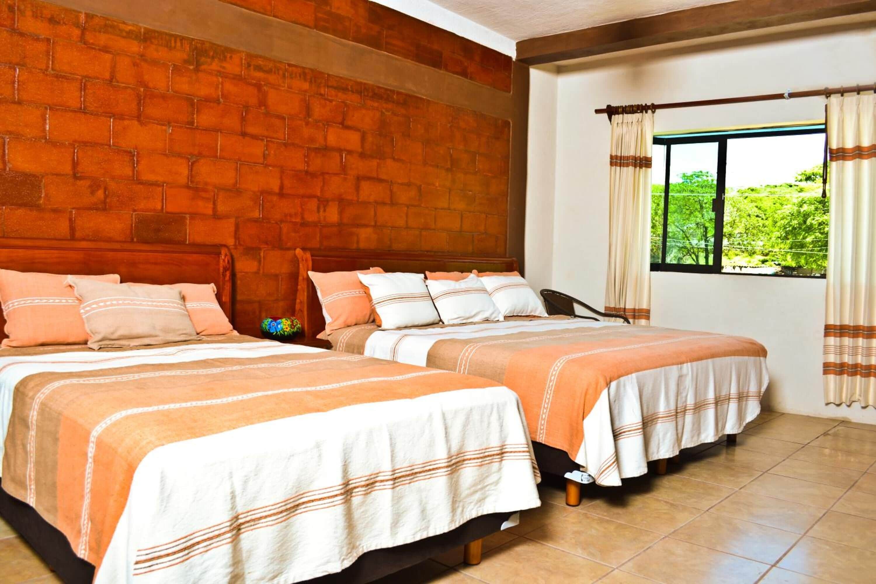 Habitación con dos camas, pared de ladrillo, ventana con cortinas y vistas al exterior con vegetación.