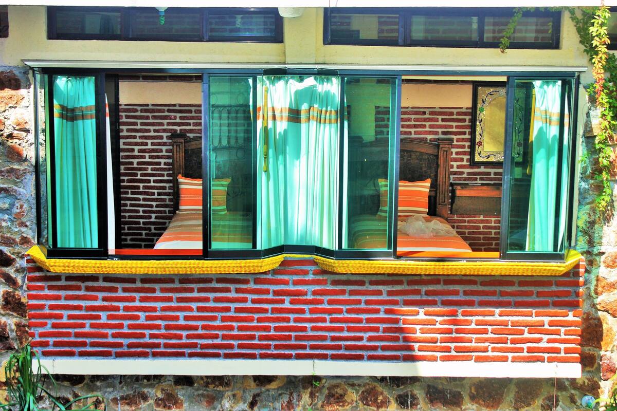 Una habitación con pared de ladrillo, una ventana con cortinas y una cama con manta naranja.