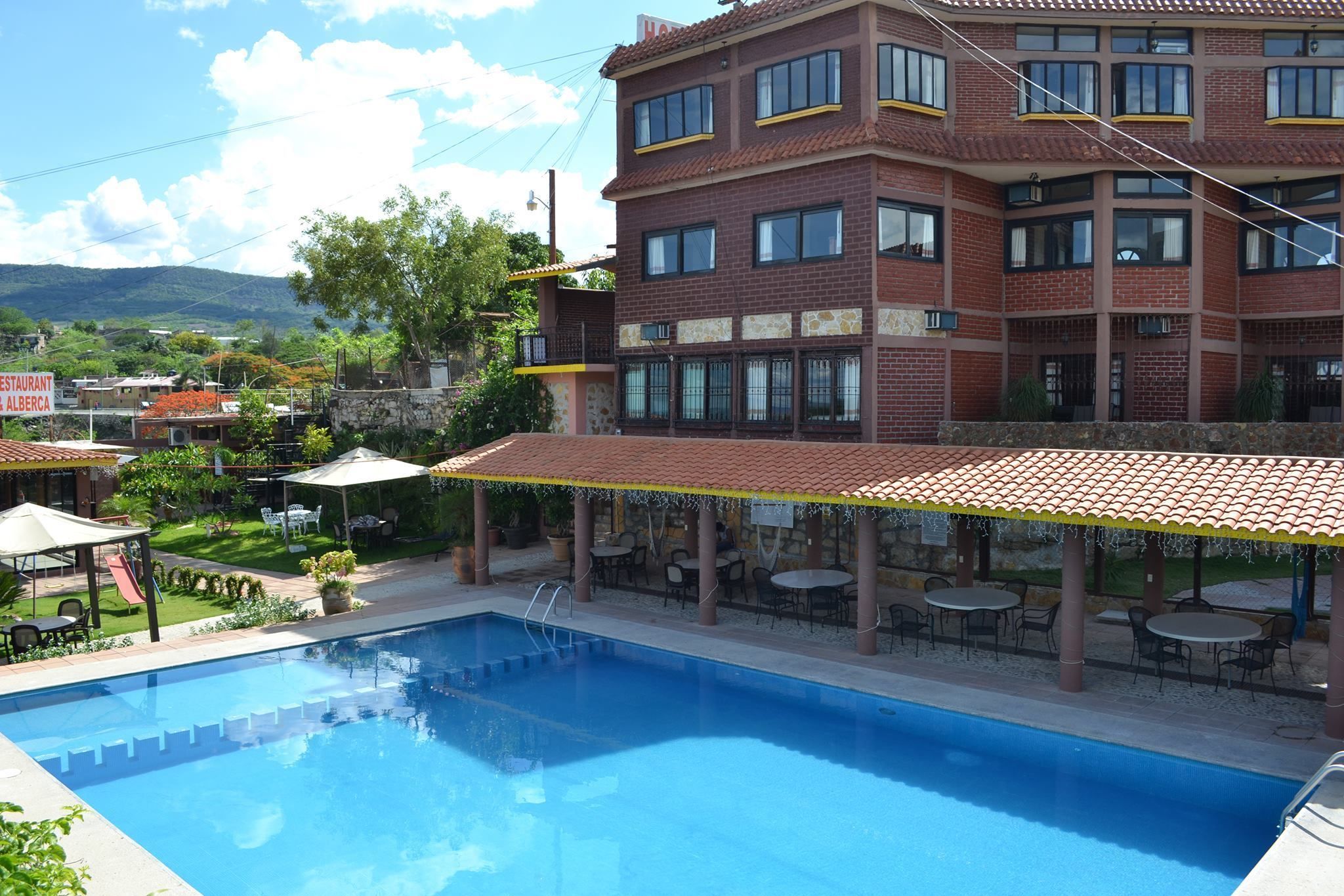 Un área de piscina con terraza cubierta, mobiliario exterior y vistas a un edificio de ladrillo con múltiples ventanas.