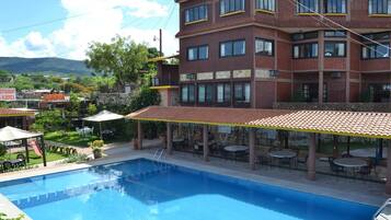 Un área de piscina con terraza cubierta, mobiliario exterior y vistas a un edificio de ladrillo con múltiples ventanas.
