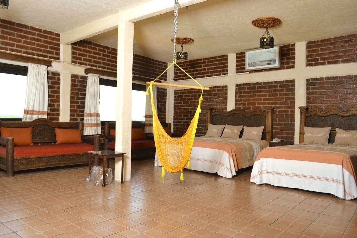 Una habitación con dos camas, un sillón hamaca, un sofá de madera y una pared de ladrillo.