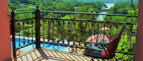 A balcony with a view of a river and greenery, a pool, and a chair.