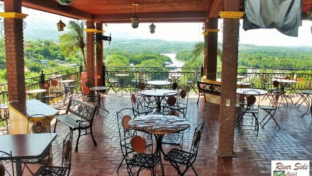 A covered outdoor seating area with tables and chairs, overlooking a lush green landscape and a river. - River Side Hotel (Chiapa de Corzo)