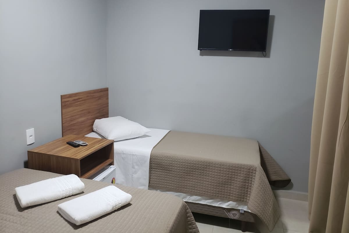 Une petite chambre d’hôtel avec deux lits, une tête de lit en bois, un téléviseur fixé au mur et une table de chevet avec une télécommande.